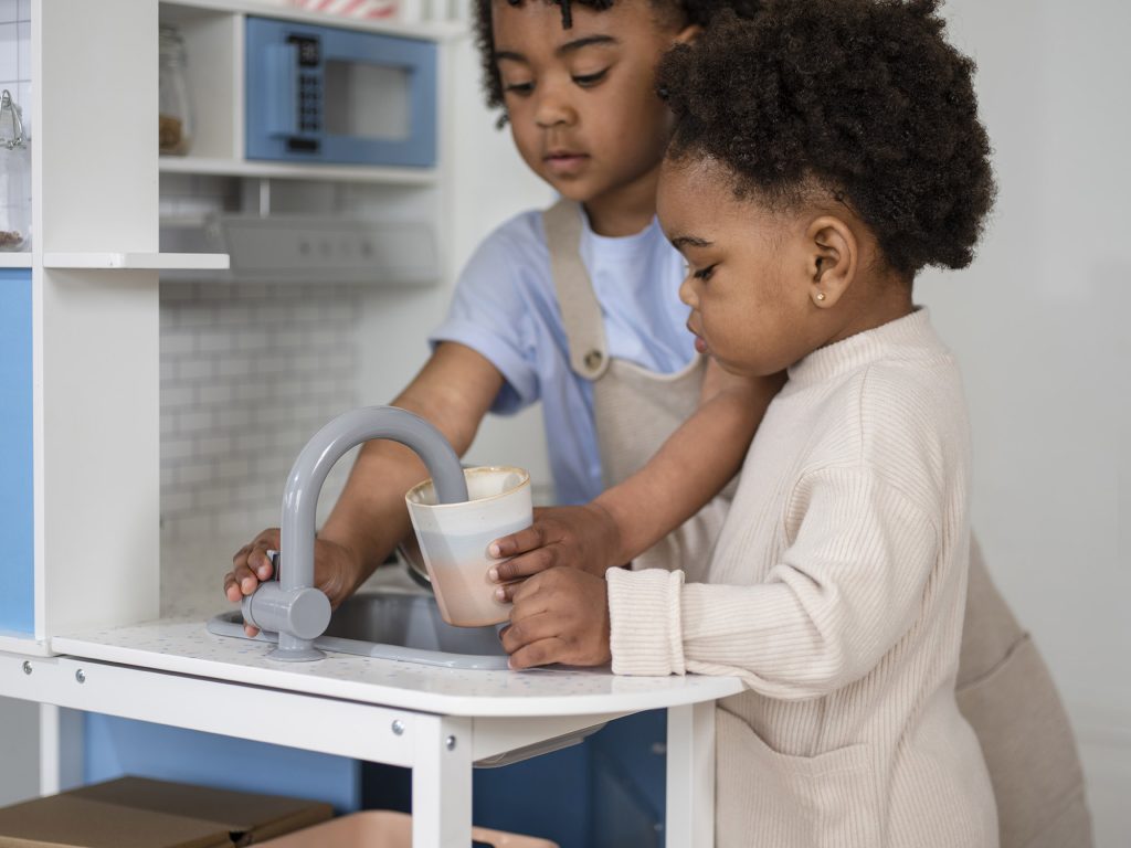 Children pretending to use taps in the Plum Penne Pantry Wooden Corner Kitchen with Fridge 